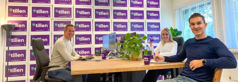 Drie mensen aan een tafel in een kantoor van Binnen Makelaars met een muur vol "Tillen"-dozen op de achtergrond.