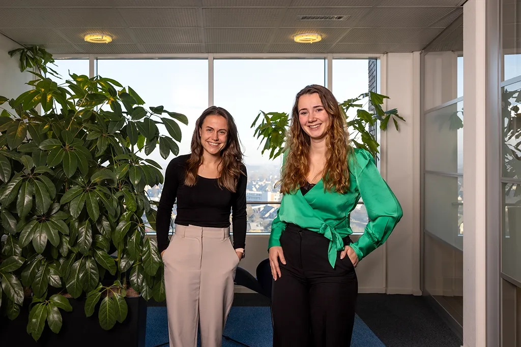 Twee vrouwen staan lachend naast elkaar bij groot raam met planten.