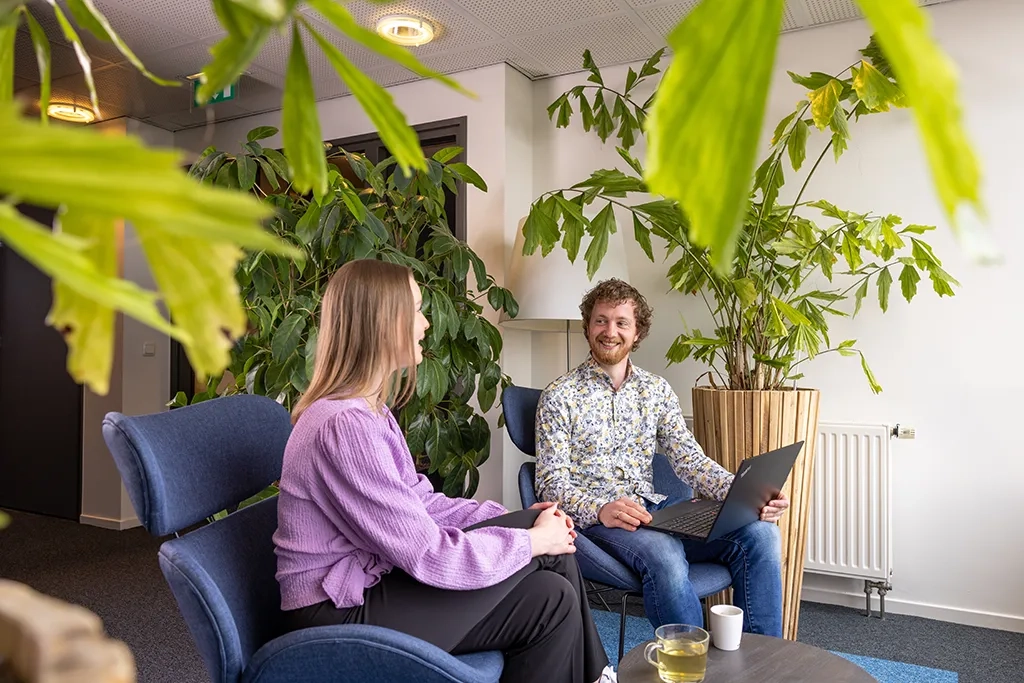 Man en vrouw praten samen in zitgedeelte met planten, man heeft laptop.