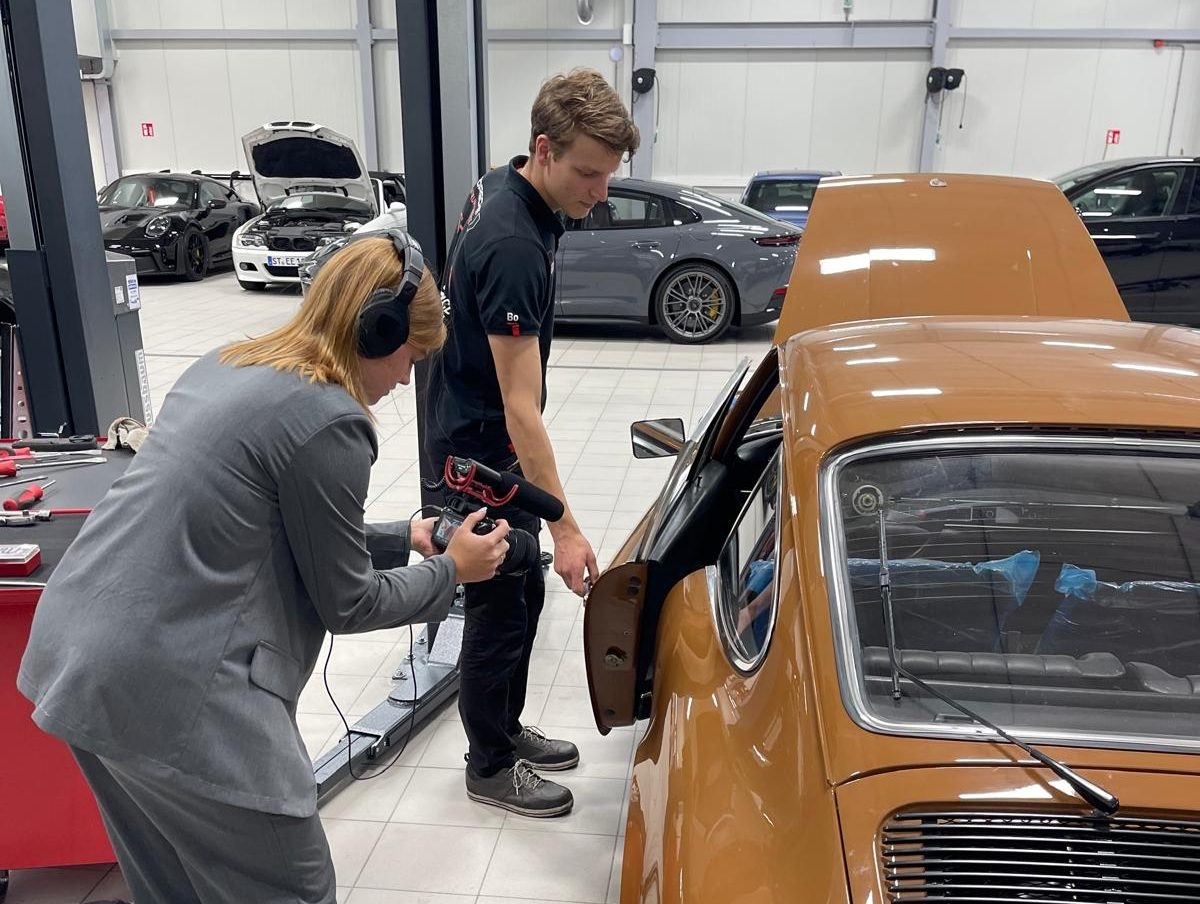 Vrouw filmt monteur die aan een klassieke Porsche werkt in een werkplaats.
