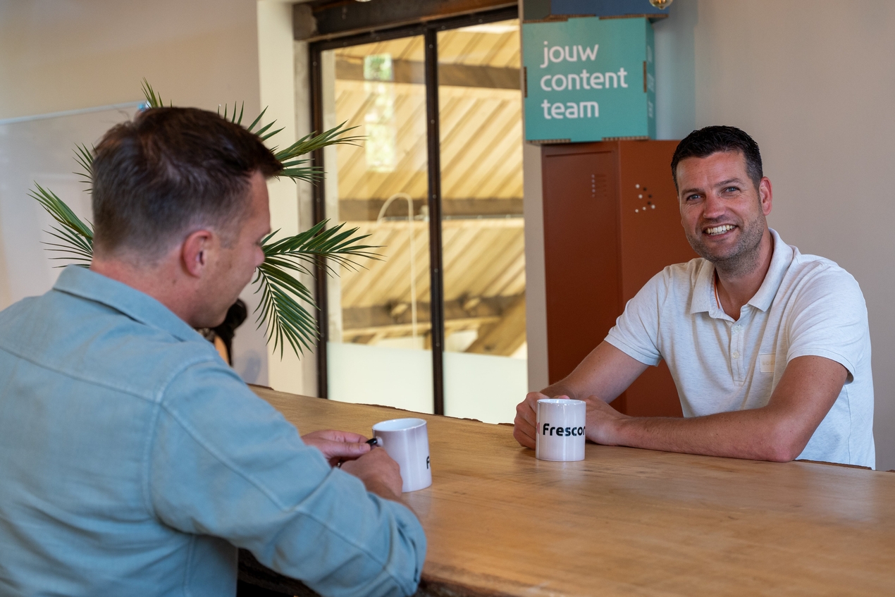 Lachende Job met Frescon-mok aan tafel in kantoorruimte.