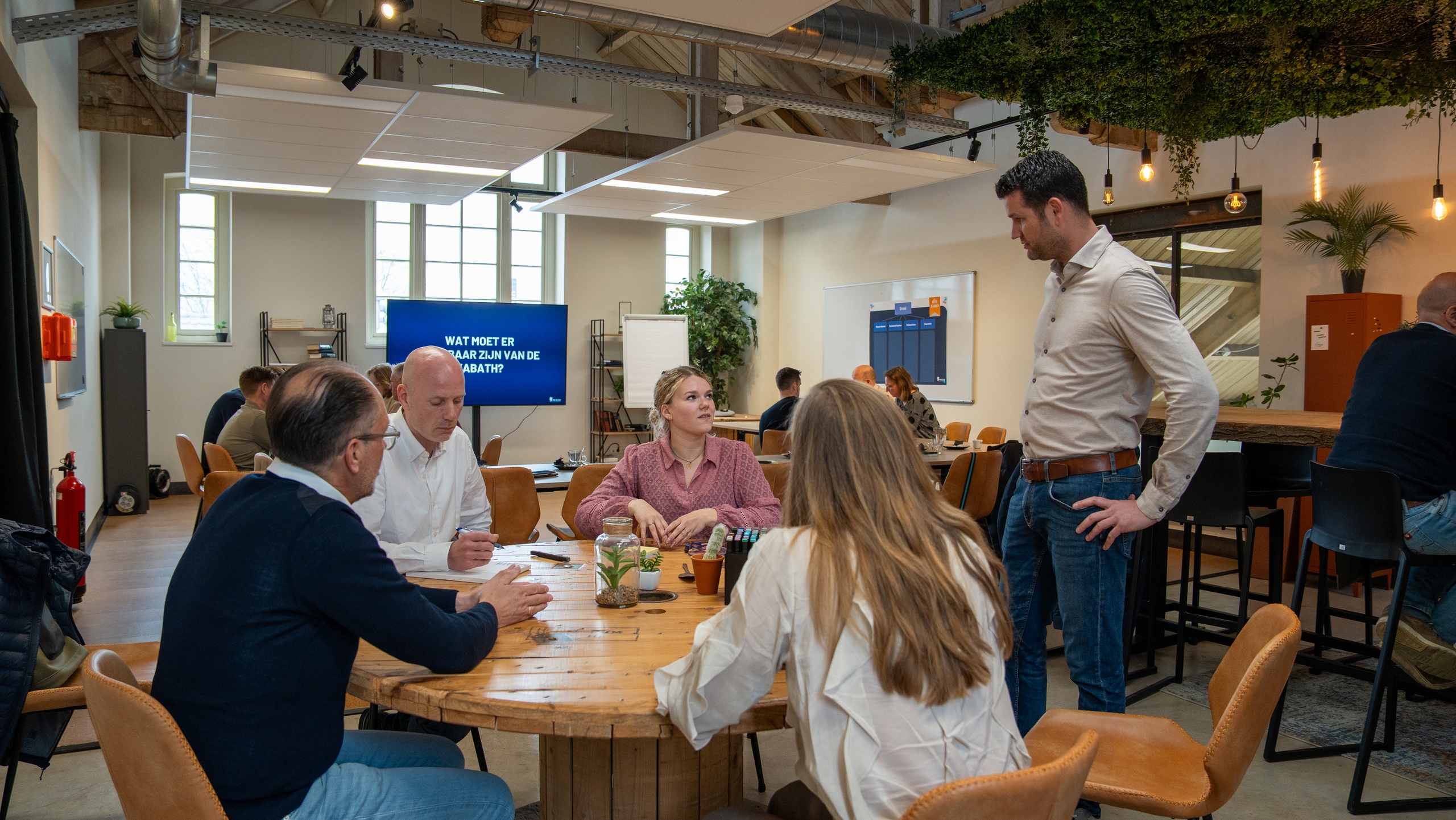 Groep mensen van de Kabath Vastgoedbeheer overlegt rond tafels in een moderne vergaderruimte met scherm en planten.