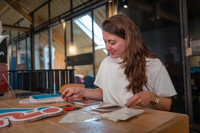 Amy zit aan een houten tafel en werkt met kleurrijke drukwerkvoorbeelden en stickers, terwijl ze geconcentreerd verschillende ontwerpen bekijkt en sorteert.
