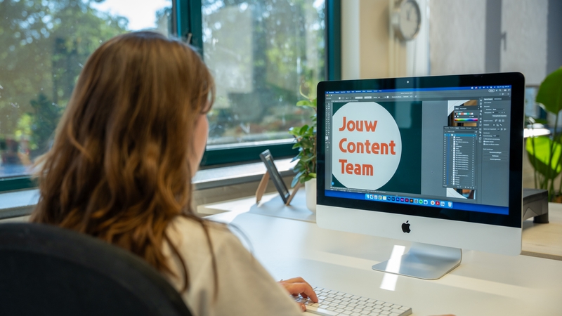 Dit is Amy, zij zit achter haar bureau en werkt op een computer aan een ontwerp waarin de tekst “Jouw Content Team” groot in beeld staat.
